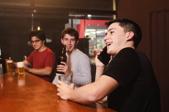 A happy young man talking on the phone while his friends enjoy a beer and laugh at the bar.