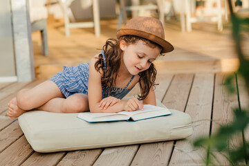 Mädchen sitzt auf Polster auf Holzterasse und liest ein Buch