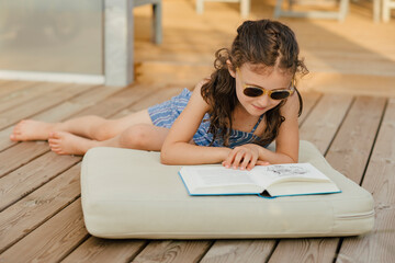 Mädchen mit Sonnenbrille sitzt auf Polster auf Holzterasse und liest ein Buch