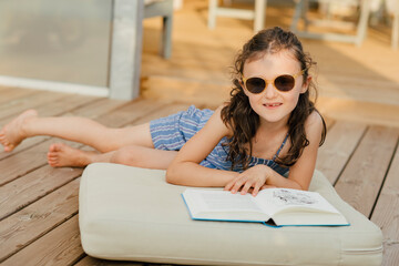 Mädchen mit Sonnenbrille sitzt auf Polster auf Holzterasse und liest ein Buch