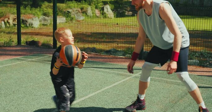 Basketball player father training son to dribble on basketball court outdoors in summertime. Sportive happy family