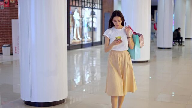 Young confused woman using real time navigation app on smartphone to find luxury clothing boutique in shopping mall while making purchases during seasonal sale, carrying many paper bags in hand