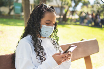 Teen girl with curly hair sitting on a park bench texting on her cell phone sitting on a bench