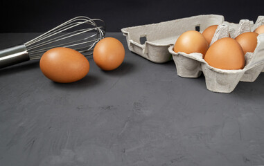 Fresh eggs in a cardboard egg tray with a whisk on a dark background