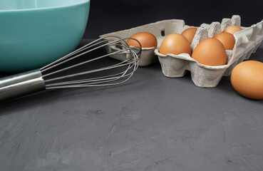 Fresh eggs in cardboard egg tray with whisk and plate and on dark background