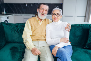 Obraz premium happy cheerful 60s couple hugging sitting on sofa in cozy modern living room posing for the camera. The concept of a strong family, long life together, harmonious relationships and love