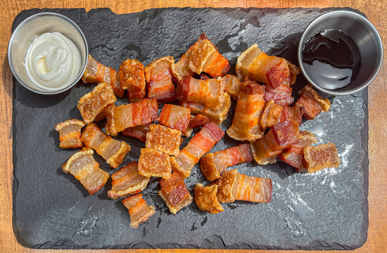 a plate of fried torreznos seen from above on a black slate plate and two jars with mayonnaise and barbecue sauces on a wooden table