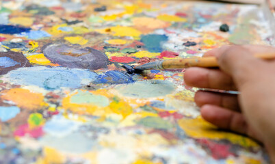 Fototapeta premium Mano tomando un pincel de pintura al oleo, en paleta de colores distintos, brillantes rojos, amarillos azules y purpuras y naranjas. Arte y artesanías creativo.