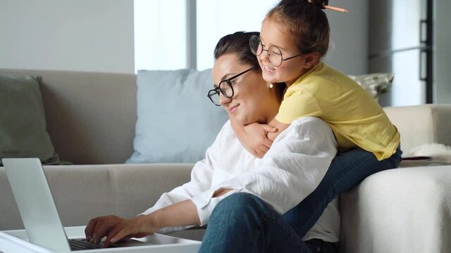 Side view of cute little girl daughter embracing her young mother working remotely from home. Mom freelance and child sitting on sofa in living room, hugging and looking at laptop screen