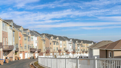 Fototapeta premium Pano Townhouses along road with white fence against scenic blue sky and white clouds