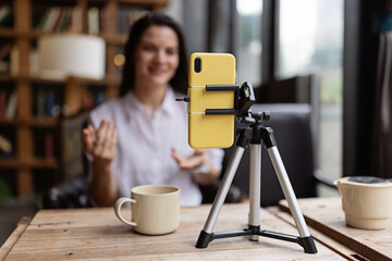 Happy young Caucasian millennial or gen z woman with long brunette hair streaming with smart phone on tripod, shooting social media blog in modern cafe. Influencer using social networks indoor. - obrazy, fototapety, plakaty