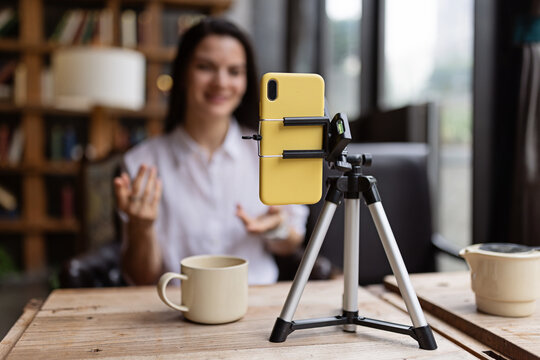 Happy young Caucasian millennial or gen z woman with long brunette hair streaming with smart phone on tripod, shooting social media blog in modern cafe. Influencer using social networks indoor.