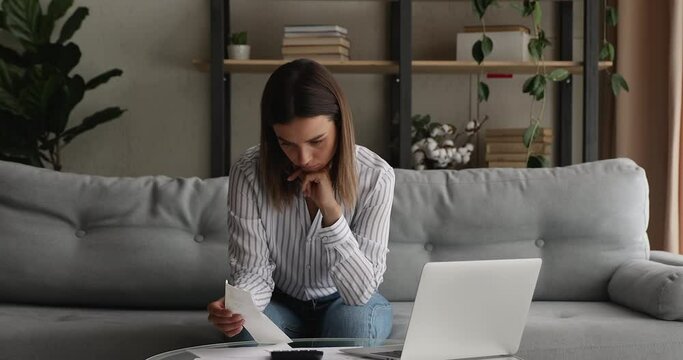 Concentrated young 20s woman calculating taxes or utility bills, making payments for household services online, involved in managing monthly budget alone at home, investment accounting concept.