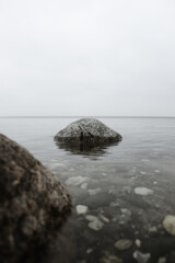Fototapeta na wymiar A large stone positioned in the sea in front of a white sky and a broad horizon. In the foreground, you can see multiple small rocks and stones on the seabed, as well as another large stone.