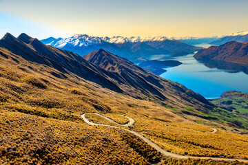 Hikers tramping  the winding path to the top of Roys Peak overlooking the lake and snow capped...