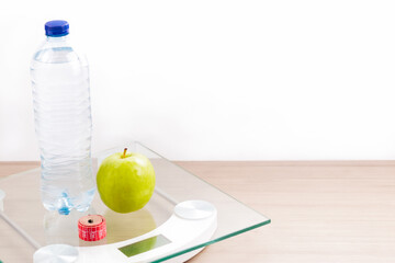 Front view of scale on wooden table with bottle of water, tape measure, apple and white background.