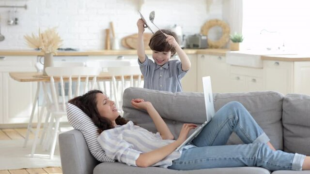 Young mother tired of small son distracting making noises and annoying as helpless woman try to rest with laptop use device for shopping online or relax watch video lying on couch at home after work