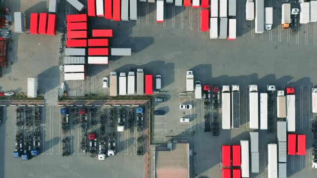 Aerial top down view of many cargo containers and semi-trailer trucks standing on a parking lot in a logistics park with warehouses and a loading hub