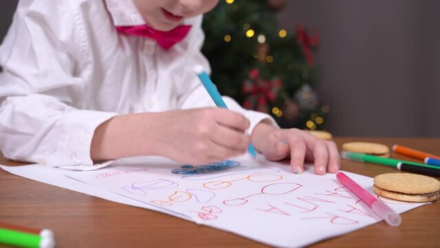 Child learns how to write. Good boy draws greeting card for his parents. Kid writes wish letter to Santa on Christmas Eve, blurred decorated Christmas tree on background.