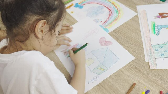 Adorable asian kid girl is drawing and coloring beautiful and colorful pictures (rainbow, house) with her mother hug shows concept of spending parental leisure time activity with happy family.
