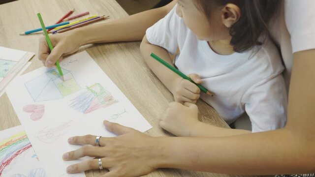 Adorable asian kid girl is drawing and coloring beautiful and colorful pictures (rainbow, house) with her mother hug shows concept of spending parental leisure time activity with happy family.