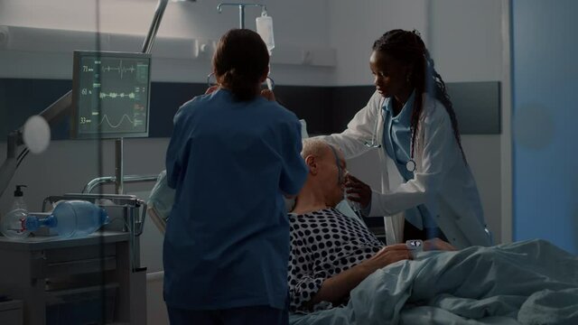 Patient with breathing problems in hospital ward bed needing reanimation help from nurse and african american doctor using oxygen tube. Old man with disease in intensive care room