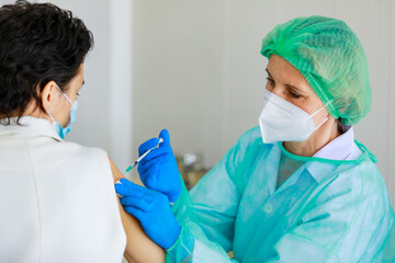 Caucasian senior female doctor in PPE full hazard protection uniform with face mask using syringe needle inject vaccine to woman patient shoulder at hospital vaccinating desk