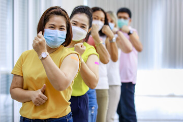 Close up shot of Asian happy strong female patient wears face mask stand look at camera in front of vaccinated orderly queue line in blurred background showing thumb up and bicep muscle up together - obrazy, fototapety, plakaty