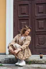 Young smiling millennial woman with wild hair dressed in an autumn coat sitting near the door of an old building with a cup of coffee to go.