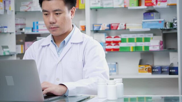 Doctor man working in pharmacy store at hospital. Asian pharmacist male looking laptop computer checking medication details in pharmacy drugstore. Concept of medical technology, business, lifestyle.