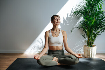 Joyful woman practicing yoga, meditating in a big studio. Frontal view. She's sitting in easy pose, looking up and to the side.