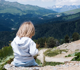 The child is sitting and looking at the landscape of the Caucasus Mountains. Outdoor travel, local travel.