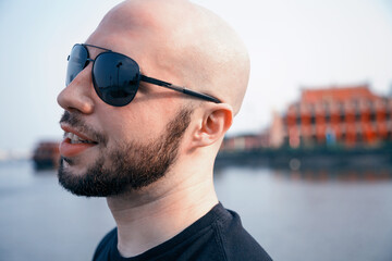 Happy young man in sunglasses strolling along a river and daydreaming. Bald bearded masculine guy...