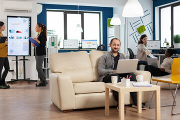 Portrait of man employee typing on laptop looking at camera smiling while diverse team working in background. Multiethnic coworkers talking about startup financial company in modern business office