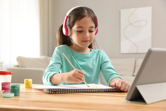 Little girl with headphones drawing on paper at online lesson indoors. Distance learning