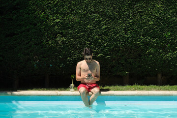 Young man wearing sunglasses using his smartphone sitting on the swimming pool edge. Summer concept.