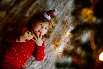 Little cute toddler girl in bed under Christmas tree, eating reindeer cookie and dreaming of Santa...