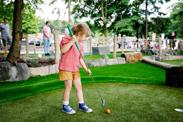 Cute preschool girl playing mini golf with family. Happy toddler child having fun with outdoor...