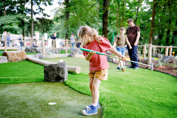 Fototapeta premium Cute preschool girl playing mini golf with family. Happy toddler child having fun with outdoor activity. Summer sport for children and adults, outdoors. Family vacations or resort.