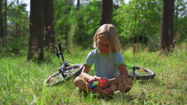 Blonde Boy plays Simple Dimple Rainbow Colors. The boy sits in the park, a bicycle lies in the back.