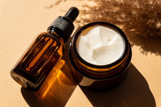 Close Up Of Collagen Cream In Glass Jar, Serum In Amber Glass Bottle And Dry Flowers Reeds On Beige Background. Set For Skin And Body Care Beauty Products