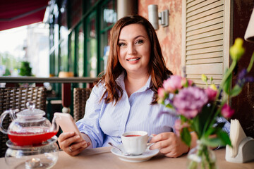 An attractive woman in a blue dress drinks cafe tea and texts in a smartphone