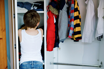 School kid boy standing by wardrobe with clothes. Child making decision for school jeans pants to...