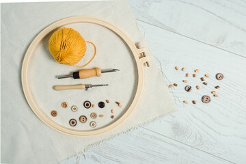 Embriodery hoop with two punch needles, ball of yellow yarn, wooden buttons and piece of linnen fabric, with some wooden buttons on white wooden floor. Punch work creative hobby.