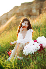 Cute girl with wavy hair wearing a hat holding a glass of wine in her hand covering part of her face Summertime. Picnic at sunset in the mountains
