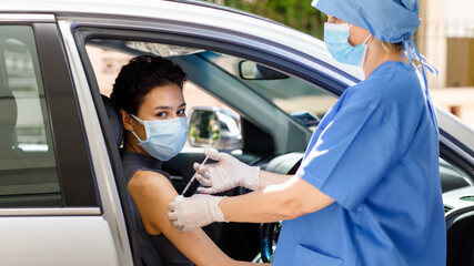 Caucasian female doctor from public health in blue hospital uniform and face mask stand hold vaccine syringe needle injection on woman patient shoulder in drive through car vaccination queue