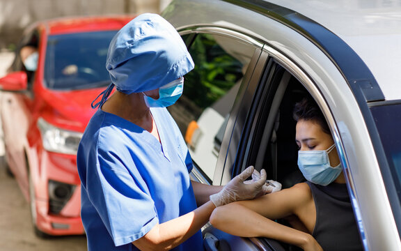 Female doctor in blue hospital uniform and face mask stand near car holding syringe in hands wears rubber gloves injecting covid 19 vaccine to drive through patient shoulder on vaccination queue