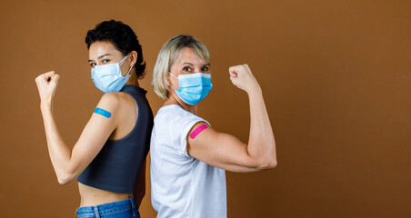 Two gender female citizens wears face mask standing look at camera lean on each other back hold fist up showing strong muscle with colorful plaster together after vaccination - obrazy, fototapety, plakaty