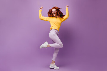 Red-haired woman in stylish pants and sweater is dancing and shouting joyfully on lilac background