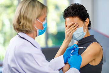 Female wears face mask sit hold hand close eye afraid of pain from vaccination while doctor using...